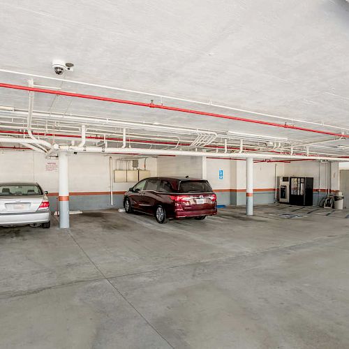 An indoor parking garage with two cars parked, a silver and a red one, and a vending machine next to the wall.