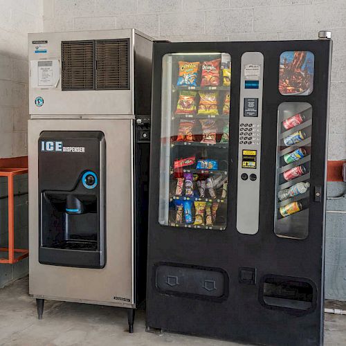 The image shows an ice dispenser machine next to a vending machine filled with various snacks and drinks, placed against a light gray wall.