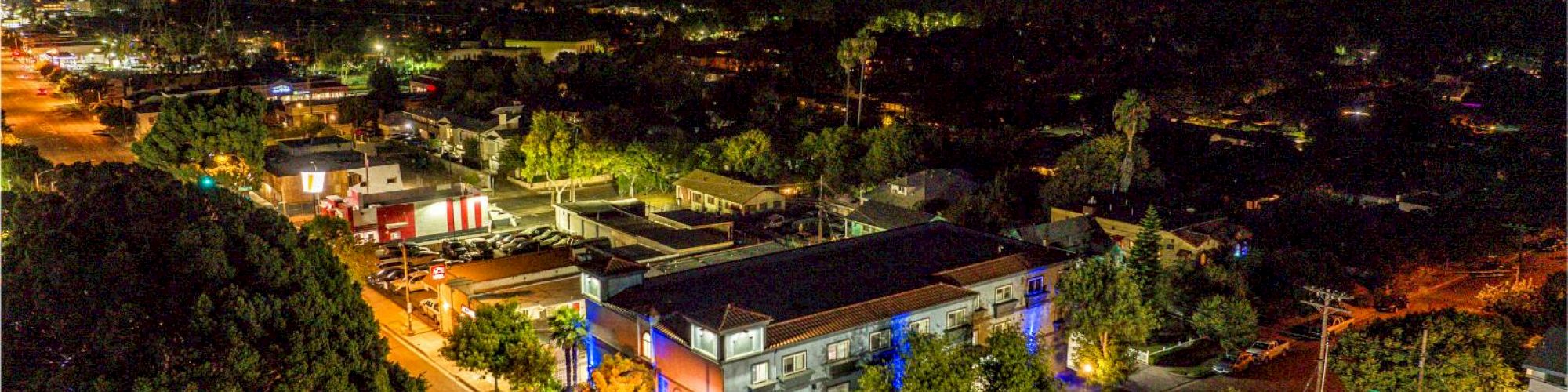 An aerial view of a brightly lit urban area at night, with streets, buildings, and parked cars visible in the image.