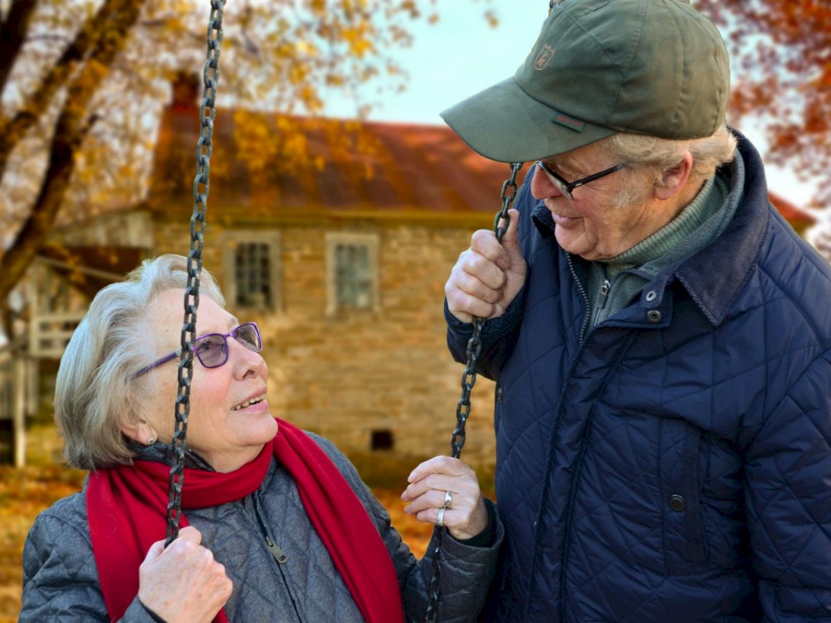 An elderly couple stands together outdoors, smiling at each other while holding onto a swing. There's a house in the background with autumn leaves.