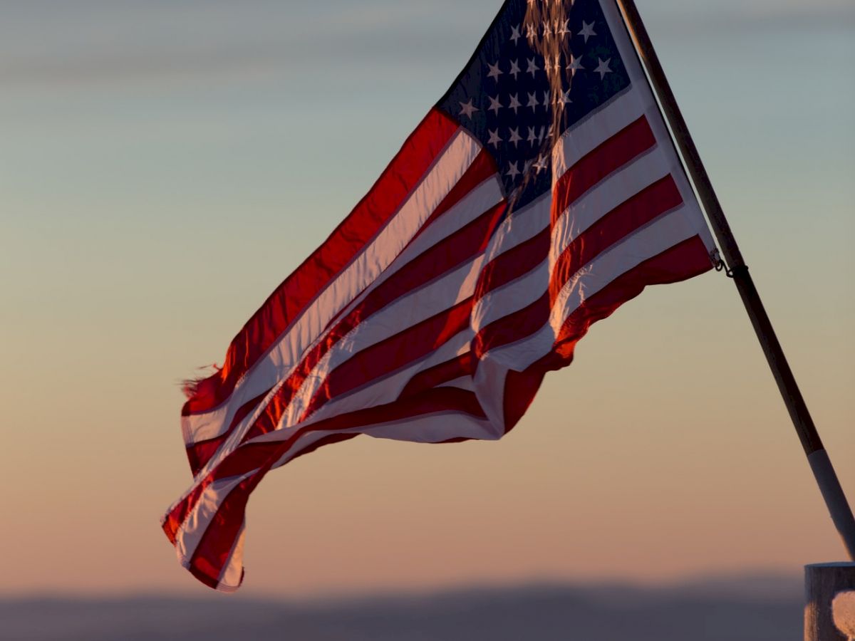 An American flag waves in the wind against a background of a colorful sky, suggesting either sunset or sunrise.