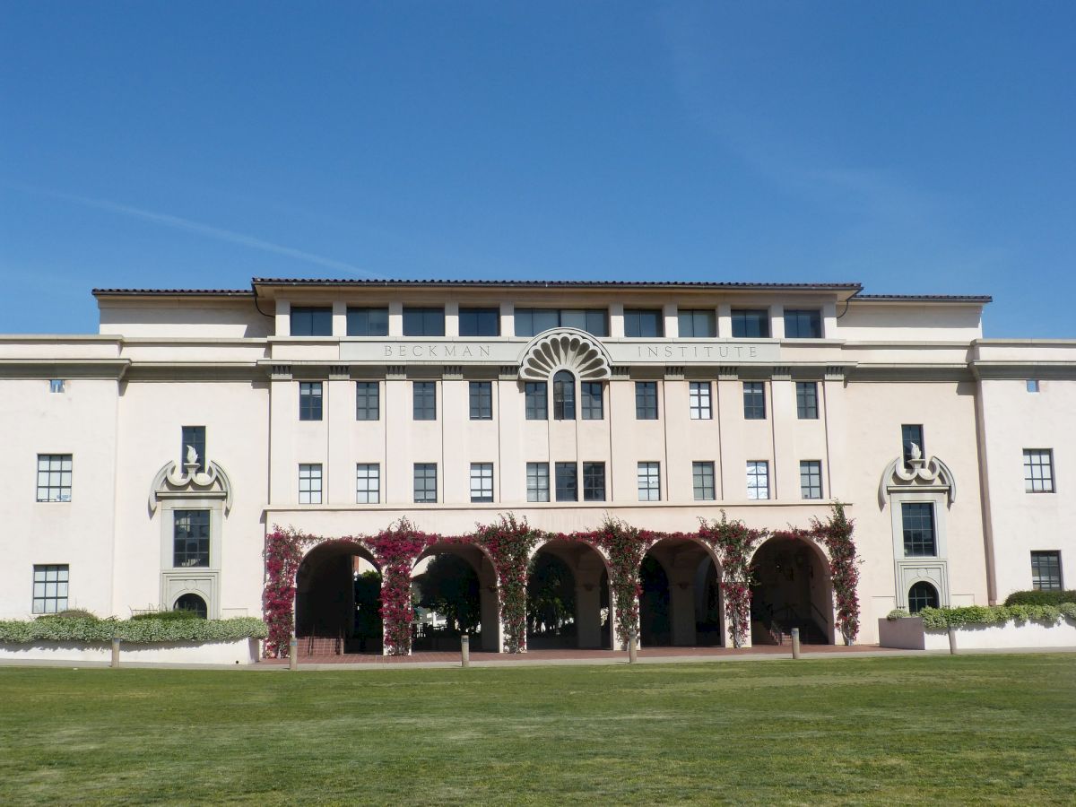 The image shows a large, multi-story building with arched entrances, surrounded by a wide, grassy area, under a clear blue sky.