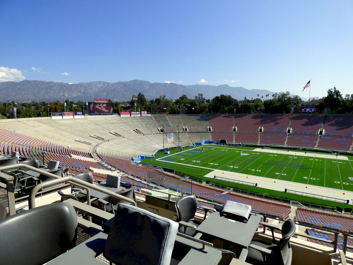 An empty sports stadium with mountains in the background, featuring green grass, seating areas, and tables in the foreground, under a clear sky.