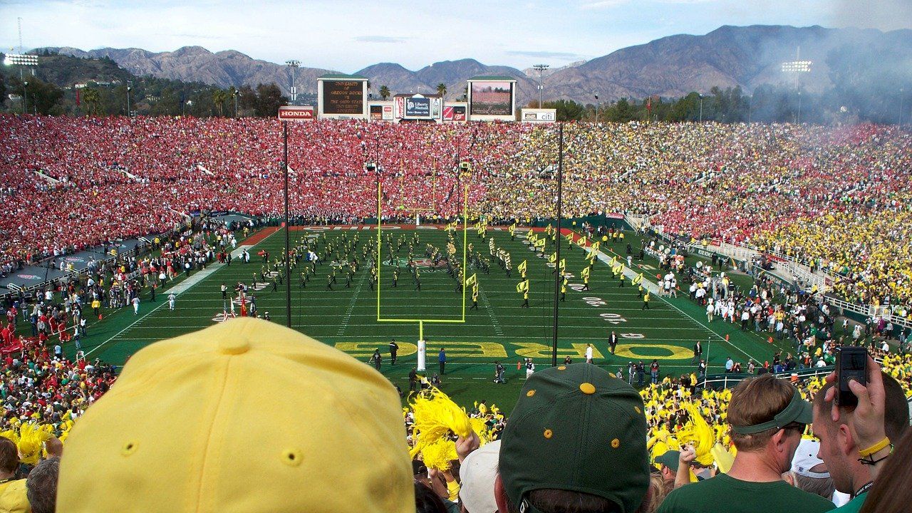 A crowded football stadium with fans wearing yellow and green on one side and red on the other, overlooking a game in progress on the field.