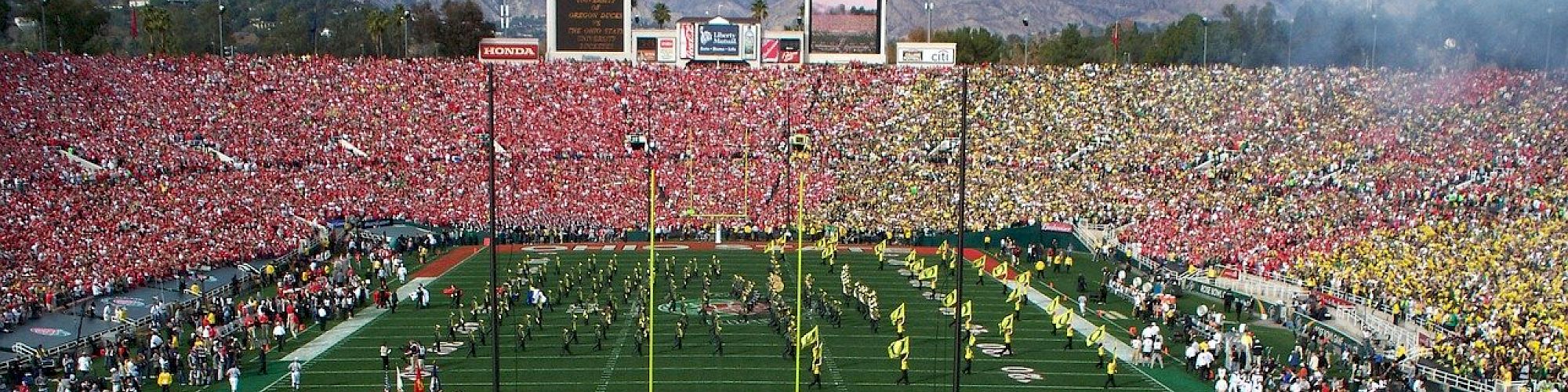 A crowded football stadium with fans wearing yellow and green on one side and red on the other, overlooking a game in progress on the field.