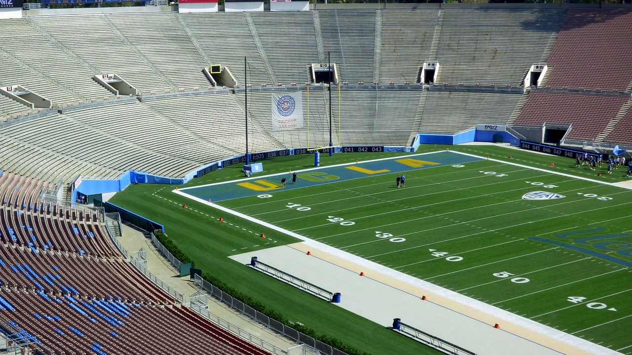 An empty football stadium with green turf, marked yard lines, and surrounding bleachers. A large screen displays a message in the distance.