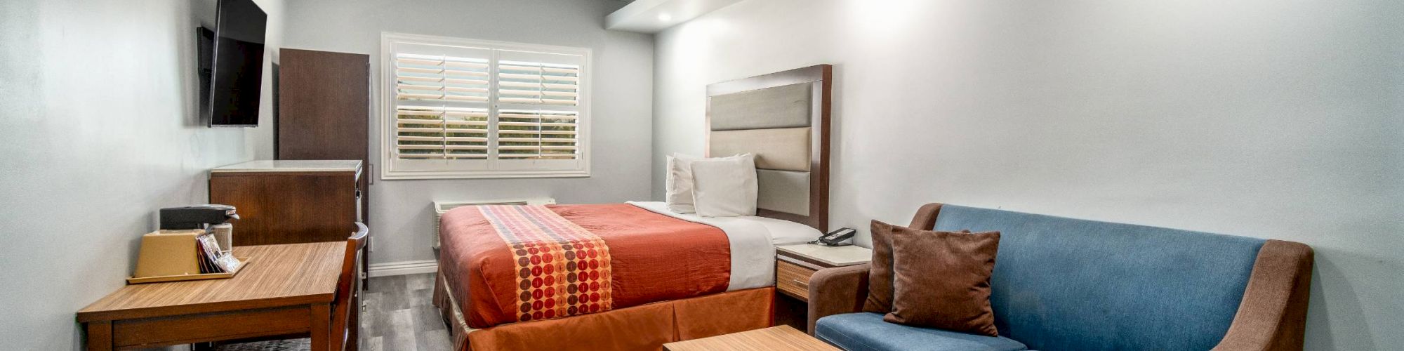 A modern hotel room with a bed, sofa, coffee table, desk, TV, and wooden floor, featuring simple white walls and ceiling lights.