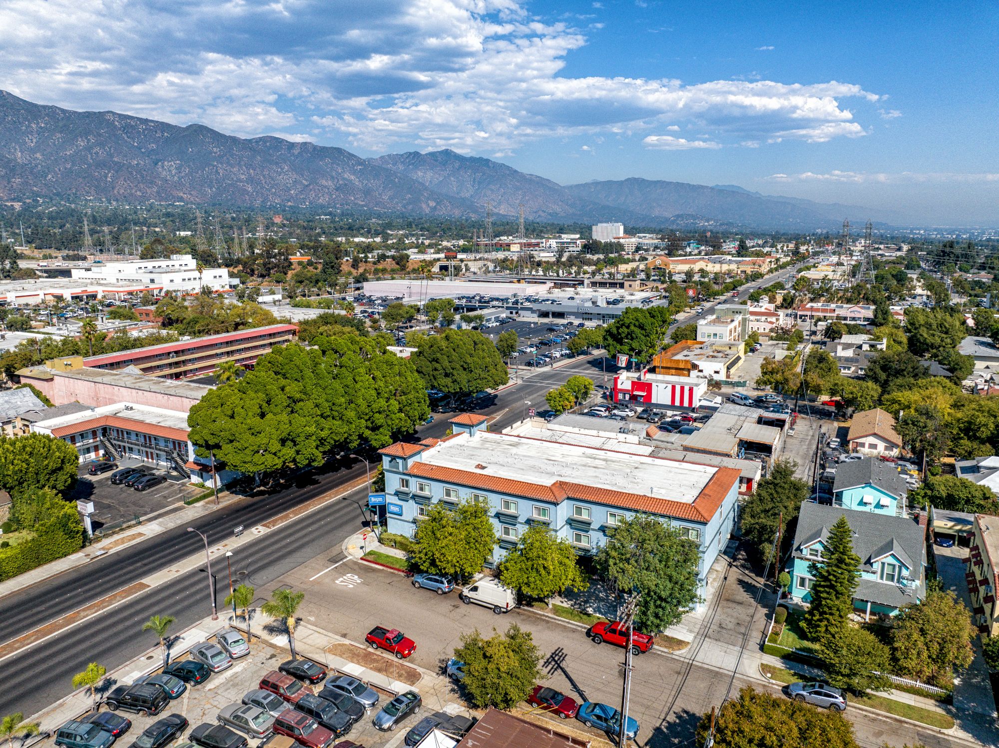 Aerial view of an urban area with streets, buildings, parked cars, trees, and distant mountains under a partly cloudy sky.