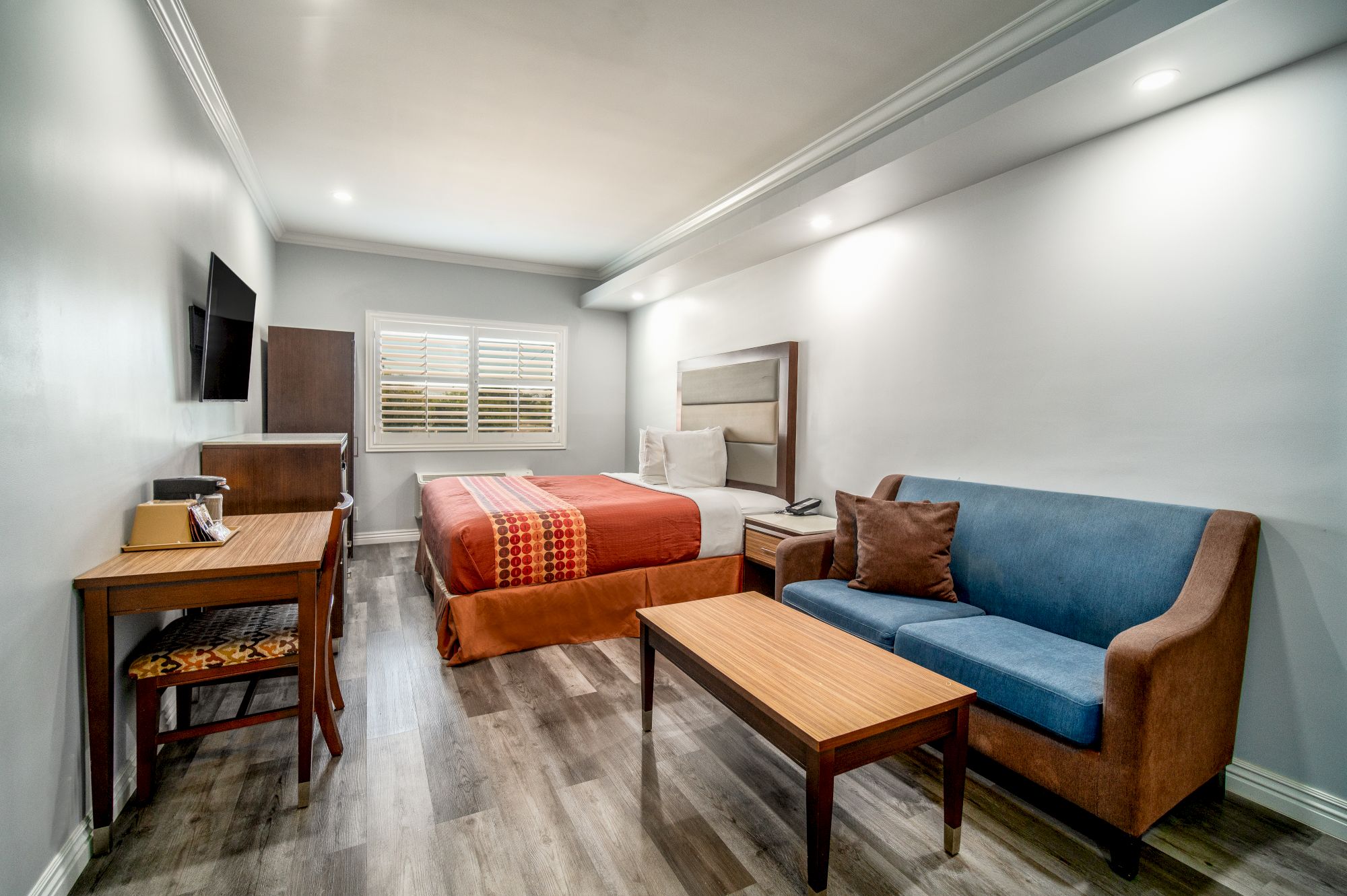 A well-lit room with a bed, two small tables, a blue sofa, wall TV, and wooden flooring.