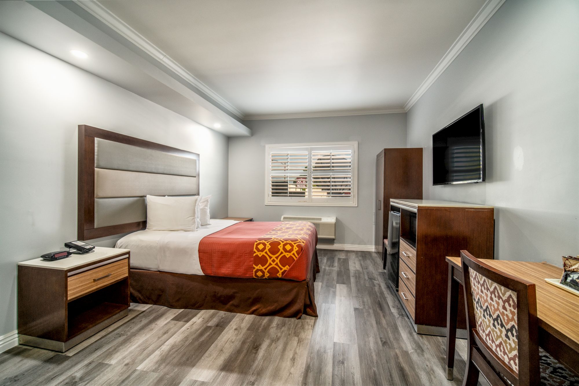 A modern hotel room with a bed, side tables, TV, and desk. The décor features wood accents and a patterned chair.