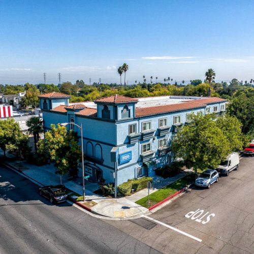 Aerial view of a blue, multi-story building at a street corner with parked cars and trees nearby on a sunny day.