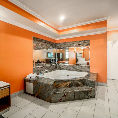 The image shows a room with a jacuzzi set against an orange wall and a large mirror, featuring modern lighting and tiling.