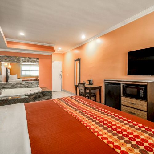 The image shows a hotel room with a bed, flat-screen TV, Jacuzzi, and a small workspace against an orange-toned wall.