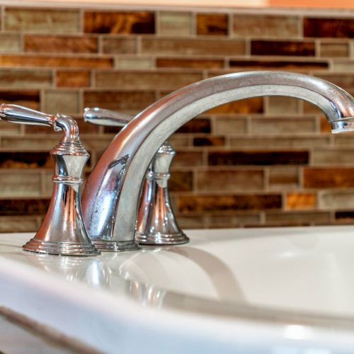 A shiny chrome faucet with dual handles is mounted on a white sink, set against a brown-tiled backsplash.