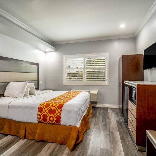 A modern hotel room with a bed, TV, wooden furniture, and a window with shutters, featuring a beige and orange color scheme.