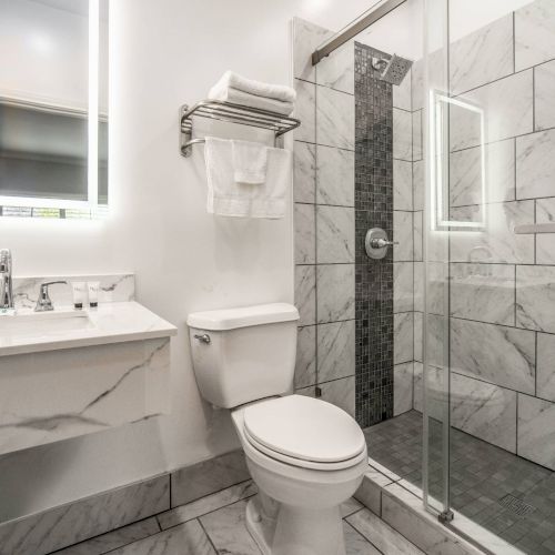 A modern bathroom with a glass shower, white tiles, a sink with toiletries, and a toilet, accented by a bright, illuminated mirror.