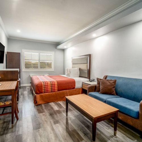 A modern hotel room with a bed, sofa, TV, desk, and table in a well-lit space with wooden flooring and minimal decor.