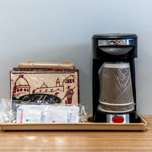 A small coffee maker on a tray with coffee supplies, including packets and a wrapped item, is displayed on a counter.