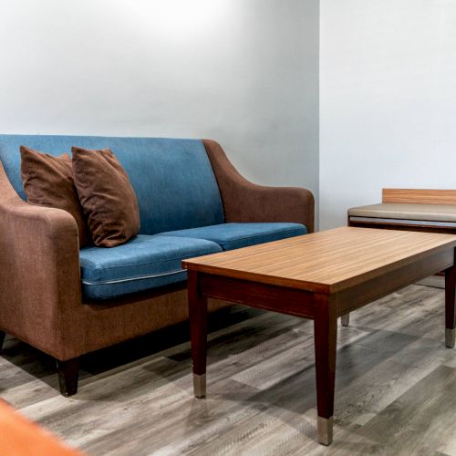 A small living room setup with a blue and brown sofa, two pillows, a wooden coffee table, and a matching side table on a wood floor.