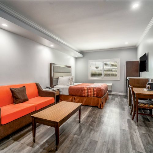 This is a modern hotel room with a bed, orange sofa, wooden table, desk, TV, and wood flooring, featuring a cozy and bright atmosphere.