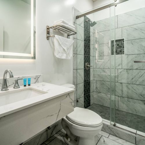 The image shows a modern bathroom with a marble countertop, sink, toilet, and a glass-enclosed shower with tiled walls and floor.