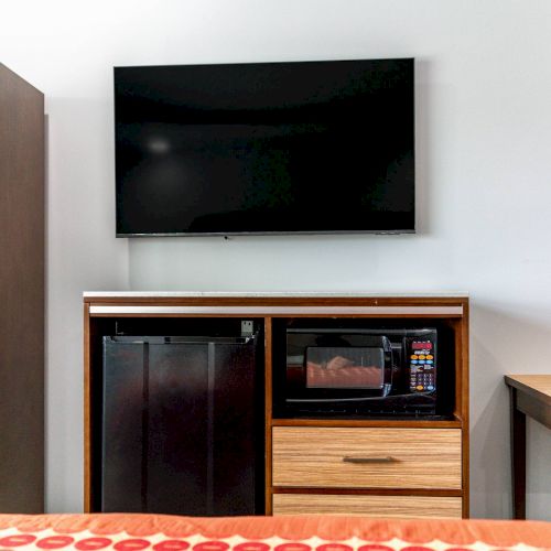 The image shows a room with a wall-mounted TV, a cabinet featuring a mini-fridge, a microwave, a wooden table, and part of a bed.