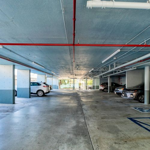 This image shows a parking garage with several parked cars, concrete pillars, and overhead pipes ending in the distance.