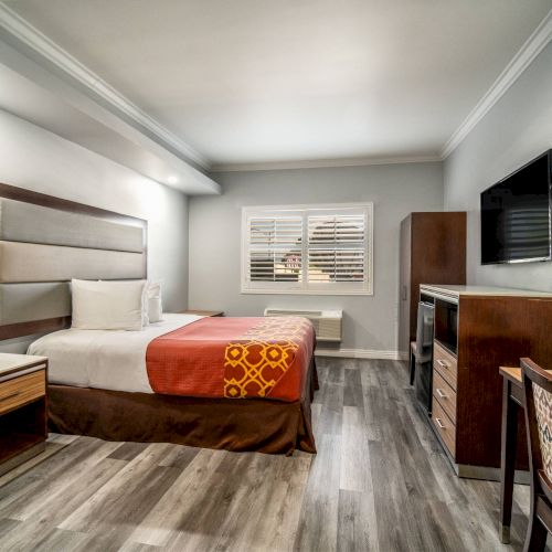 A neatly furnished hotel room with a bed, desk, TV, and cabinet. The decor is modern, featuring wooden furniture and gray flooring.