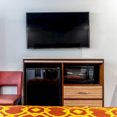 The image shows a hotel room setup with a wall-mounted TV, a mini-fridge, a microwave, a red chair, and part of a bed with a patterned cover.