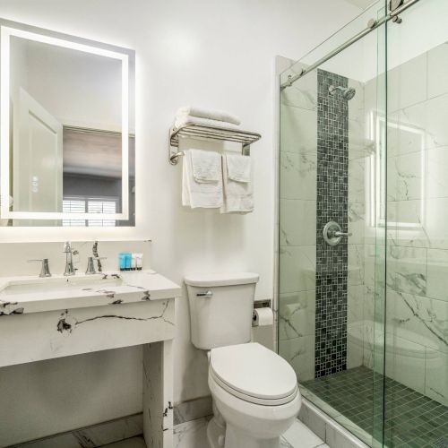 A modern bathroom with a marble countertop, illuminated mirror, toilet, and glass-door shower with tiled accents and a towel rack.