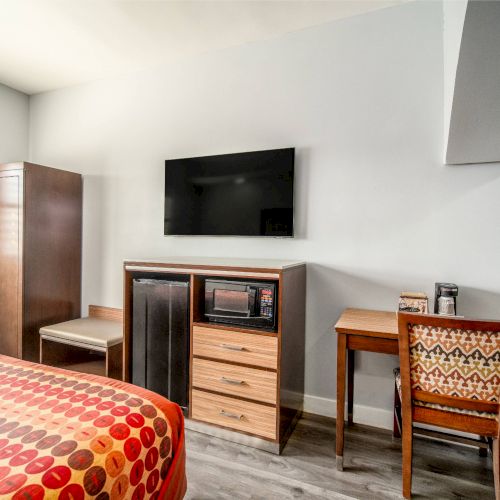 A hotel room with a bed, TV, microwave, and desk with chair; modern and minimalistic furniture setup.