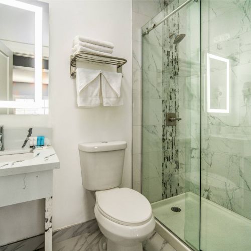 A modern bathroom with a marble shower, toilet, and sink. There are towels on a rack and a backlit mirror above the sink.