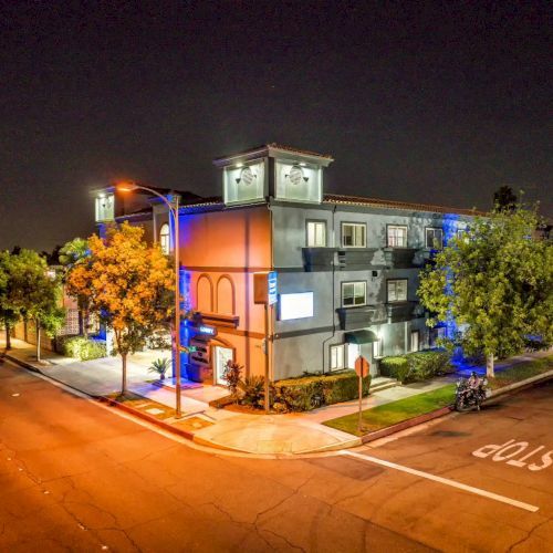 A nighttime view of a street corner featuring a lit-up building with trees lining the sidewalk and a 