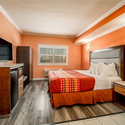 A cozy hotel room featuring a double bed with orange bedding, a wall-mounted TV, and warm lighting, in a vibrant orange interior.