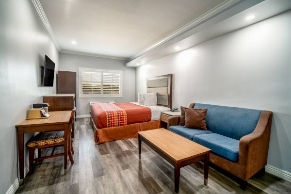 A neatly arranged hotel room with a bed, sofa, wooden table, desk, chair, and a wall-mounted TV. The decor is simple and modern.