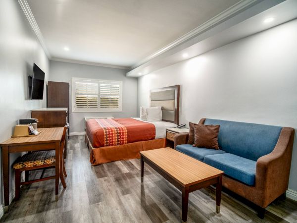 A neatly arranged hotel room with a bed, sofa, wooden table, desk, chair, and a wall-mounted TV. The decor is simple and modern.