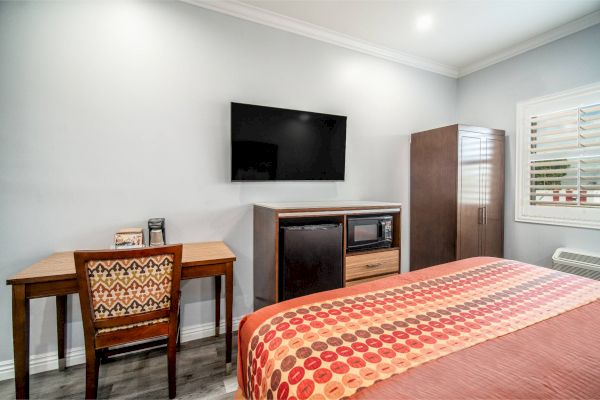 A modern room with a bed, TV, desk, and chair. A cabinet holds a microwave, mini-fridge, and storage space. Window has shutters.