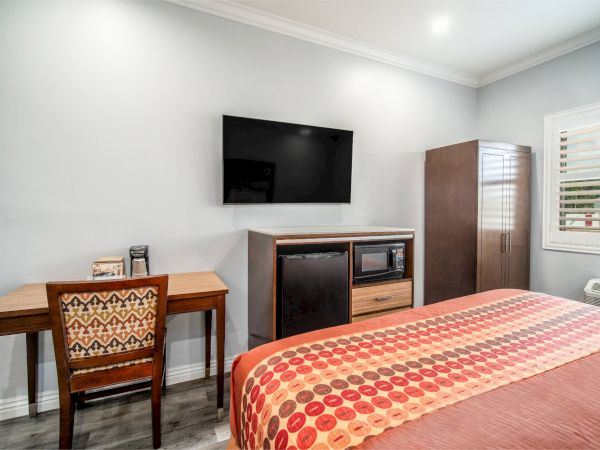 A modern room with a bed, TV, desk, and chair. A cabinet holds a microwave, mini-fridge, and storage space. Window has shutters.