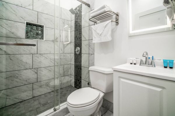 The image shows a modern bathroom with a glass-door shower, tiled walls, a toilet, a vanity with a sink, and toiletries on the counter.