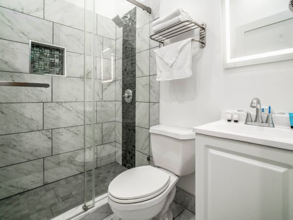 The image shows a modern bathroom with a glass-door shower, tiled walls, a toilet, a vanity with a sink, and toiletries on the counter.