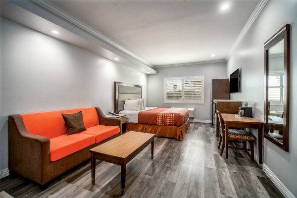 This image shows a modern hotel room with a bed, orange sofa, wooden table, TV, desk, and chair on a wood-patterned floor.