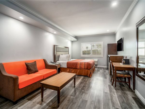 This image shows a modern hotel room with a bed, orange sofa, wooden table, TV, desk, and chair on a wood-patterned floor.
