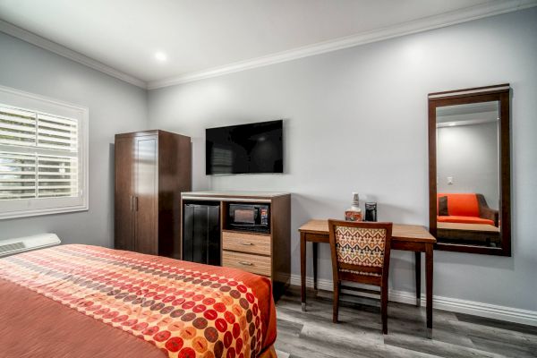 A modern hotel room with a bed, TV, desk, wardrobe, and mirror, featuring orange and red accents on the bedspread and chair.