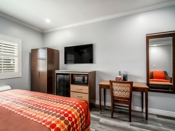 A modern hotel room with a bed, TV, desk, wardrobe, and mirror, featuring orange and red accents on the bedspread and chair.