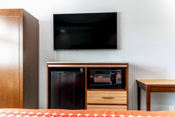 A TV mounted on a wall, a cabinet with a mini fridge and microwave, a drawer, part of a bed, and a wooden table are visible.