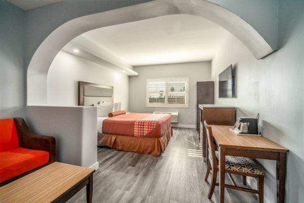 The image shows a modern hotel room with a bed, desk, chair, and sofa, all under a stylish archway, in neutral tones.