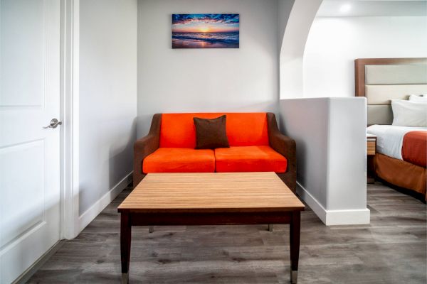 A room with an orange sofa, brown pillow, wooden table, wall art, and part of a bed visible on the side.