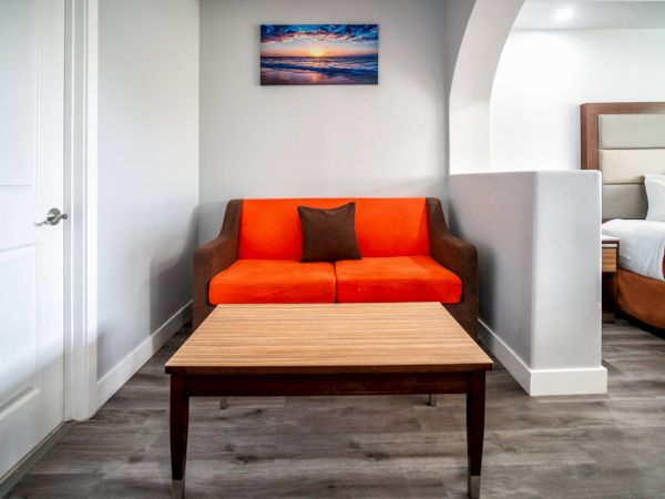 A room with an orange sofa, brown pillow, wooden table, wall art, and part of a bed visible on the side.