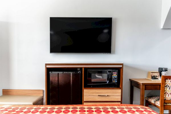 The image shows a hotel room setup with a wall-mounted TV, fridge, microwave, and a coffee maker on a counter next to a seating area.