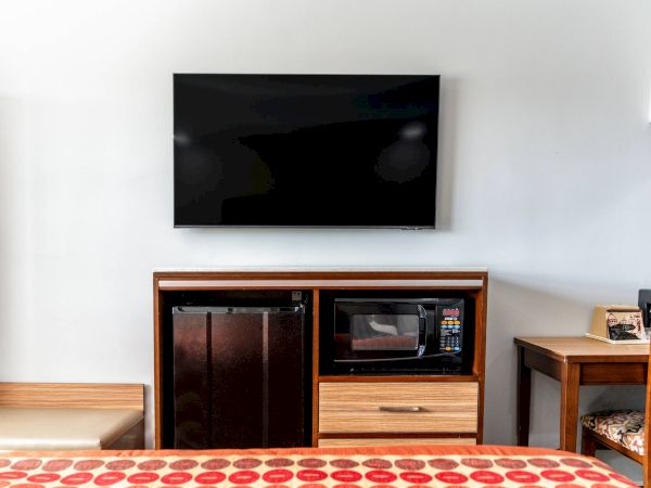 The image shows a hotel room setup with a wall-mounted TV, fridge, microwave, and a coffee maker on a counter next to a seating area.
